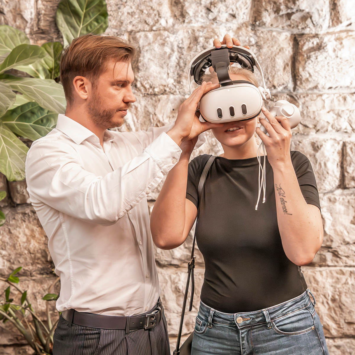 Man assisting a woman with a VR headset outdoors against a stone wall.